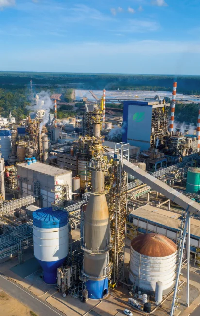 Vista aérea de uma refinaria industrial com diversas torres de processamento, tanques e tubulações sob um céu azul claro.