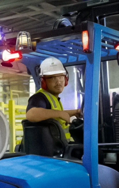 Homem usando capacete branco, protetor auricular vermelho e colete refletivo dirigindo um empilhadeira azul em ambiente industrial.