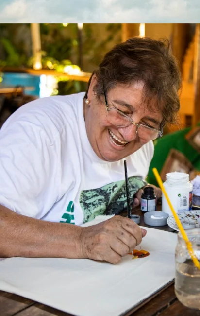 Mulher sorridente com óculos pintando em uma tela branca em uma mesa de madeira.