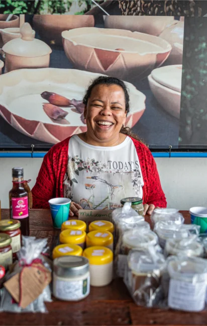 Mulher sorridente sentada atrás de uma mesa com frascos de produtos artesanais para venda.