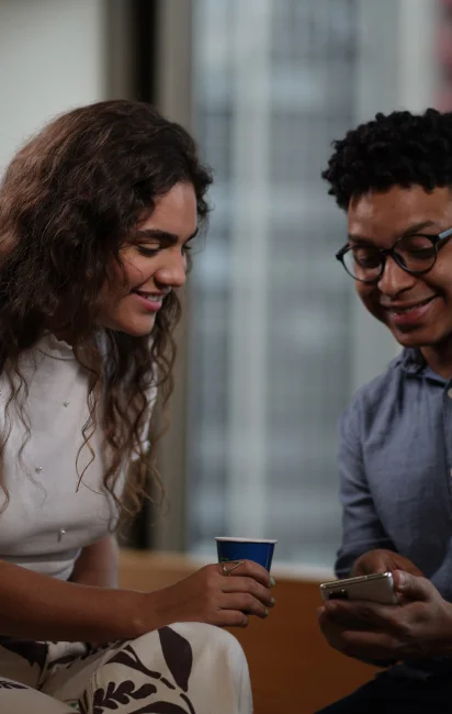 Duas pessoas sorrindo enquanto olham para um celular, uma segurando um copo azul.