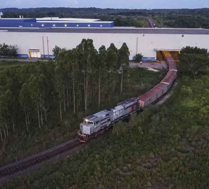 Trem de carga saindo de um grande galpão industrial cercado por vegetação e árvores.