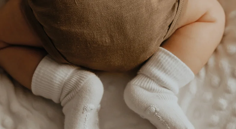 Pernas de bebê vestindo meia branca e calça marrom, deitado sobre uma superfície macia branca.