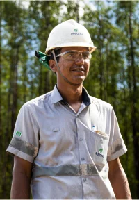Trabalhador florestal vestindo capacete branco, óculos de proteção e uniforme cinza com floresta ao fundo.