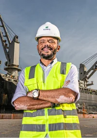 Engenheiro de obra com colete refletivo amarelo e capacete branco posando com braços cruzados em frente a um navio no porto.