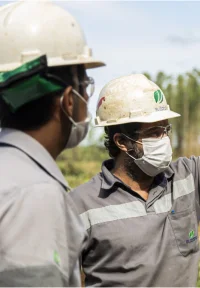 Dois trabalhadores usando capacetes e máscaras de proteção em um ambiente externo.
