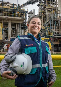 Mulher sorridente usando colete de segurança azul e segurando capacete branco em frente a uma planta industrial.