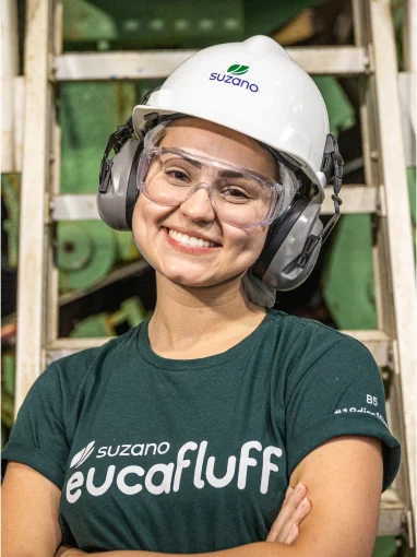 Mulher sorridente usando capacete branco �ڰ�������, óculos de proteção e abafadores de ouvido, vestindo camiseta verde com logo 'eucafluff' em ambiente industrial.