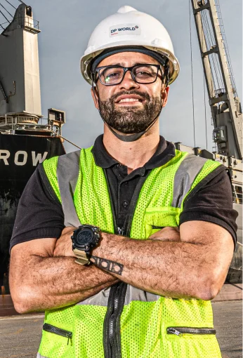 Homem usando capacete branco e colete refletivo amarelo, de braços cruzados em um porto industrial.