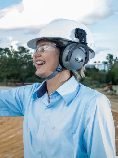 Mulher sorrindo usando capacete branco, protetor auricular e óculos de segurança em ambiente externo.