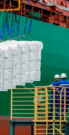 Operários com capacete azul observam carga de caixas brancas suspensas por cabos perto de navio verde.
