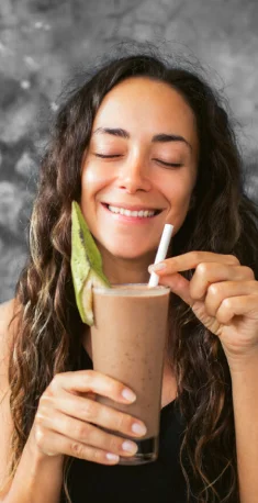 Mulher sorridente com olhos fechados segurando um copo alto com smoothie marrom e folha verde, usando canudo branco.
