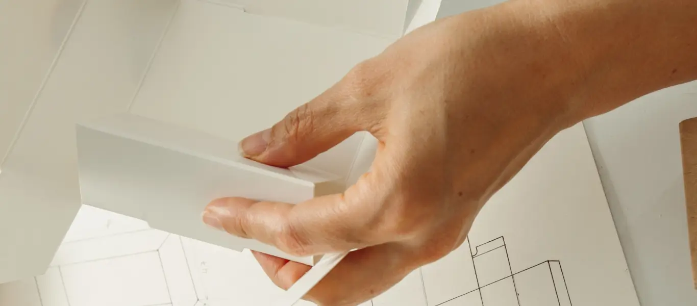 Mão segurando um modelo branco de papelão sobre um desenho técnico para montagem de embalagem.