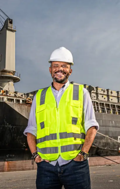 Homem sorridente usando capacete branco e colete refletivo amarelo em um cais próximo a um navio cargueiro.