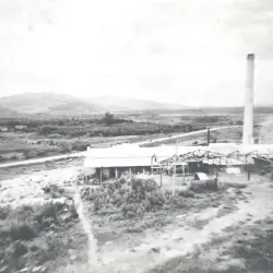 Foto em preto e branco de uma fábrica antiga com chaminé alta situada em área rural com vegetação e colinas ao fundo.