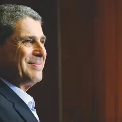 Homem de cabelo grisalho com terno escuro sorrindo de perfil com fundo desfocado marrom.