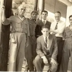 Foto em preto e branco de cinco homens, quatro em pé e um agachado, todos vestidos com roupas formais e casuais, em frente a uma parede de pedra.