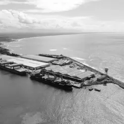 Vista aérea em preto e branco de um complexo industrial à beira-mar com vários armazéns e estruturas ao longo de um porto.