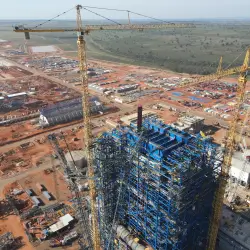 Vista aérea de canteiro de obras com estrutura metálica azul e guindastes amarelos em área rural.