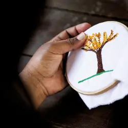 Mão segurando um bastidor de bordado com uma árvore de flores amarelas em um fundo branco.