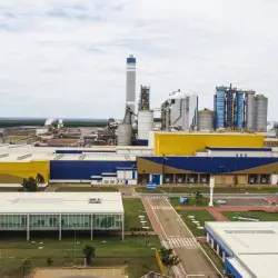 Vista aérea de uma fábrica industrial com prédios amarelos, azuis e torres altas de chaminé e silos, em um dia nublado.
