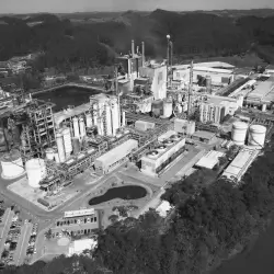 Visão aérea em preto e branco de uma fábrica industrial com várias torres, tanques e estruturas, cercada por vegetação e com um lago próximo.