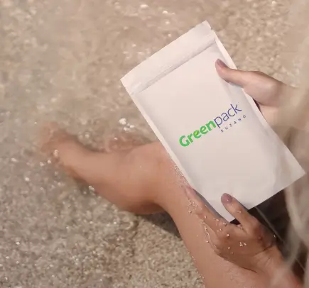 Pessoa segurando embalagem branca com logo Greenpack �ڰ������� perto de água e areia.