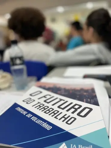 Manual do voluntário intitulado 'O Futuro do Trabalho' sobre uma mesa com pessoas desfocadas ao fundo.