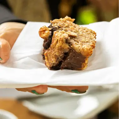 Pessoa segurando um pedaço de bolo de chocolate com nozes sobre um guardanapo branco.
