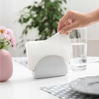Mão retirando um guardanapo branco de um porta-guardanapos cinza em uma mesa com vaso de flores, copo de água e prato.