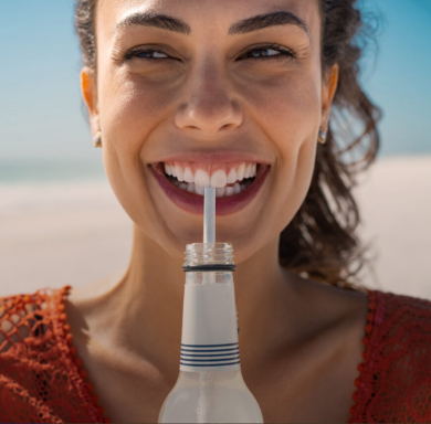 Mulher sorridente bebendo de uma garrafa com canudo em um ambiente ao ar livre.