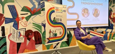 Mulher sentada em cadeira amarela numa sala colorida com um cartaz com arco-íris e palavras sobre orgulho LGBTQI+ e tela com tema de transfeminismo.