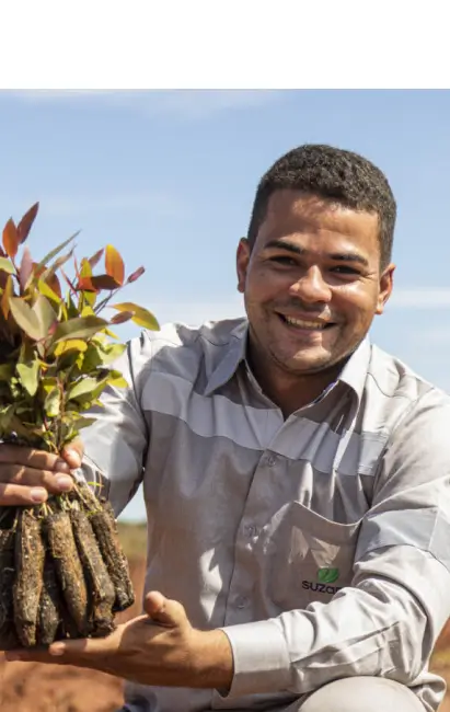 Homem sorridente segurando um conjunto de mudas de plantas com raízes expostas sob céu azul.