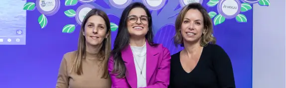Três mulheres sorridentes posando juntas diante de um fundo roxo com gráficos e ícones relacionados a iniciativas de diversidade e empreendedorismo.