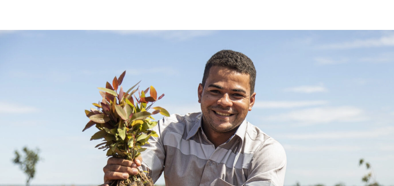 Homem sorridente segurando muda de planta com folhas verdes e vermelhas ao ar livre sob céu azul.