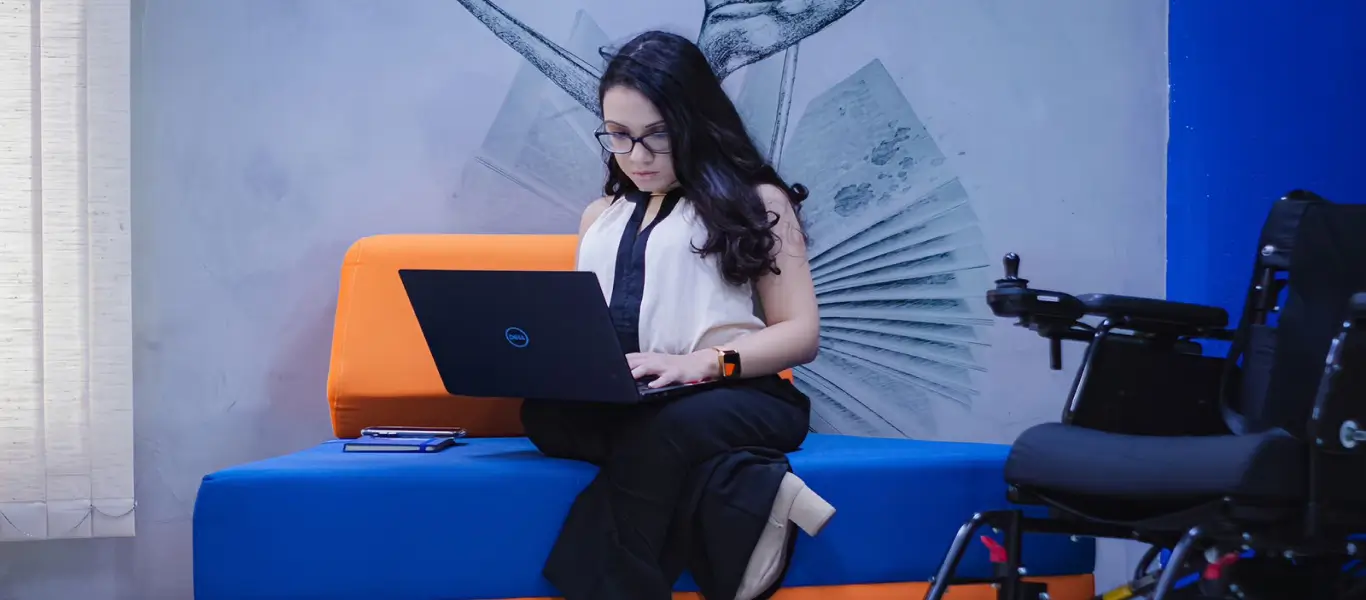 Mulher sentada em sofá azul e laranja usando laptop com cadeira de rodas ao lado.
