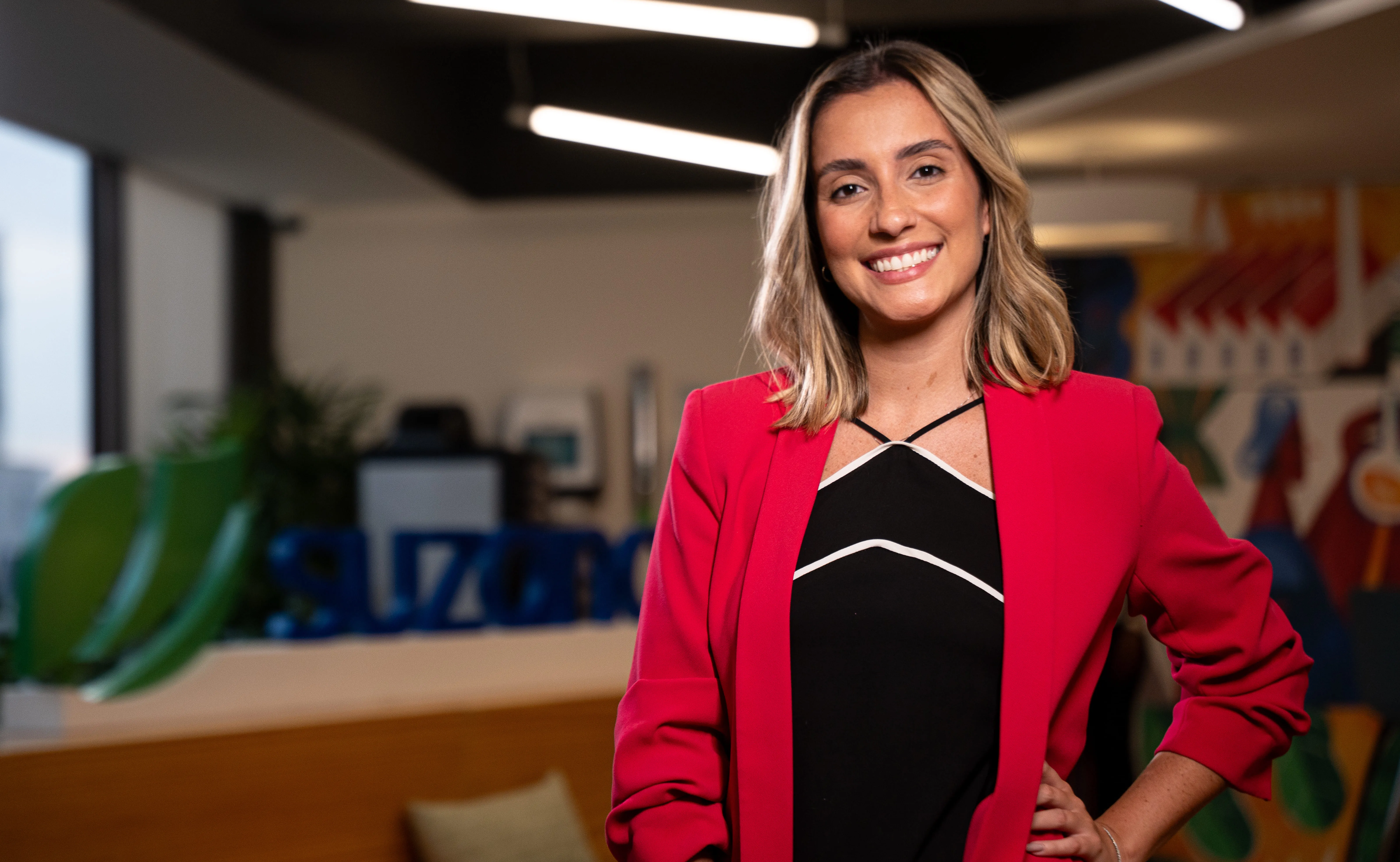 Mulher jovem sorrindo vestindo blazer vermelho em escritório moderno com decoração colorida ao fundo.