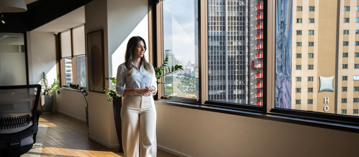 Mulher jovem vestindo camisa branca e calça clara olhando pela janela em escritório moderno com vista de prédios urbanos.