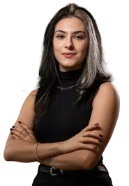 Mulher jovem com cabelo castanho e mechas loiras, vestindo blusa preta sem mangas, com braços cruzados e expressão confiante.