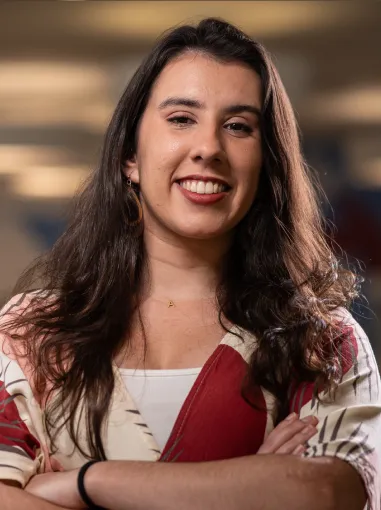 Mulher sorridente com cabelos longos e escuros, vestindo blusa branca com detalhes vermelhos e braços cruzados.