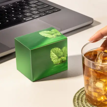 Cubinho de papel cartão verde com estampa de folhas, ao lado de um notebook e um copo de chá com gelo.