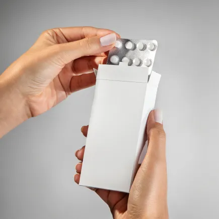 Mãos segurando uma caixa branca de medicamento enquanto retiram um blister com comprimidos redondos.