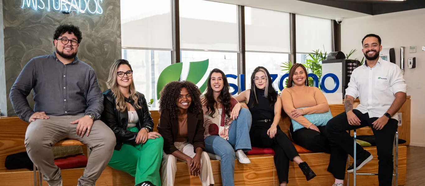 Grupo de sete jovens adultos sentados sorrindo, em ambiente de escritório moderno com logo �ڰ������� ao fundo.