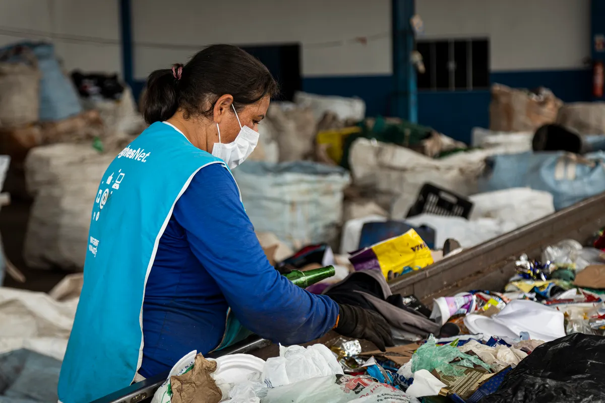 Mulher com máscara facial e colete azul separa recicláveis em uma esteira com lixo plástico e papel em centro de reciclagem.