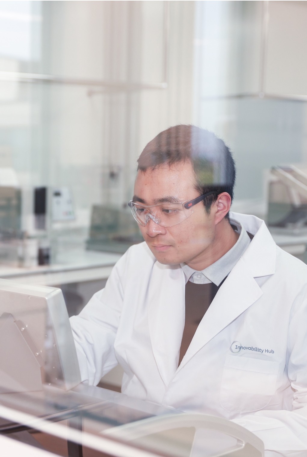Cientista usando óculos de proteção e jaleco branco trabalhando com equipamento em laboratório moderno.