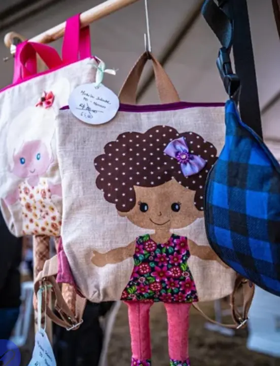 Três mochilas penduradas, duas com bonecas estilizadas e uma xadrez azul e preta.