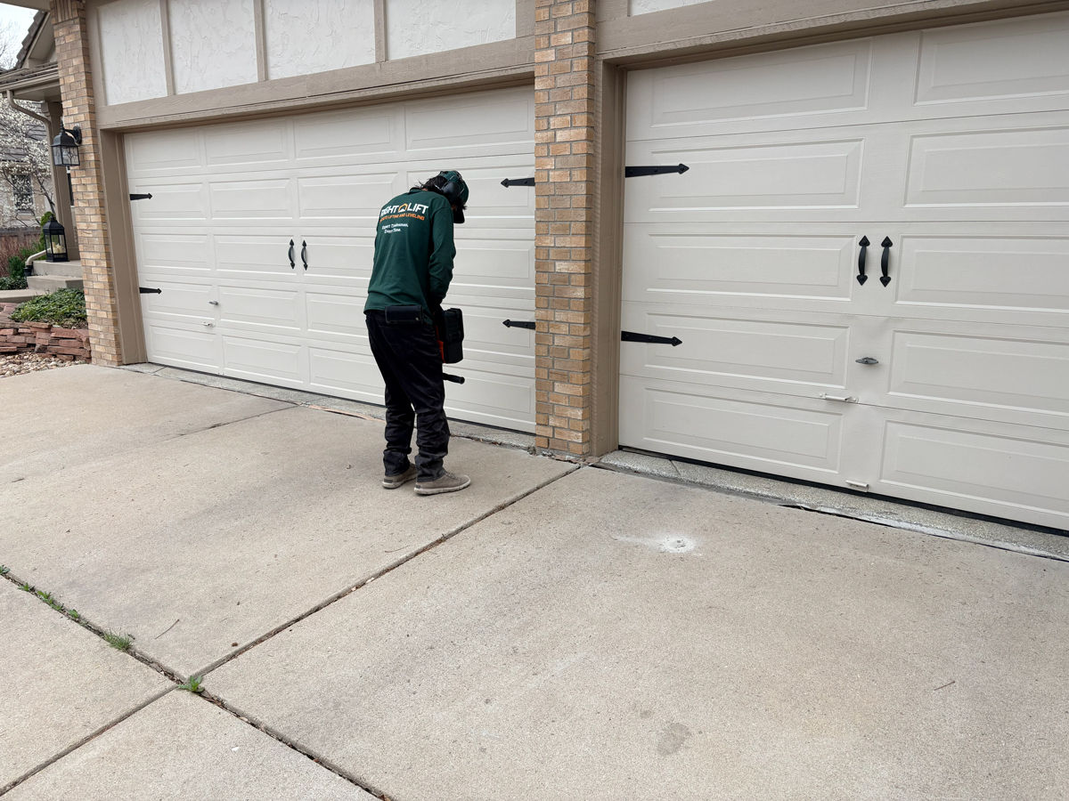 Expert tradesmen using equipment to lift and level concrete in front of a two-car garage with white doors.