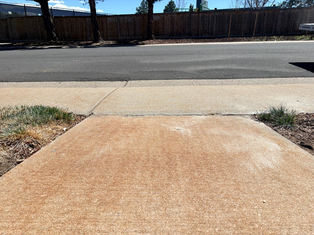 Concrete sidewalk ending at a street with a wooden fence and trees in the background under a blue sky.