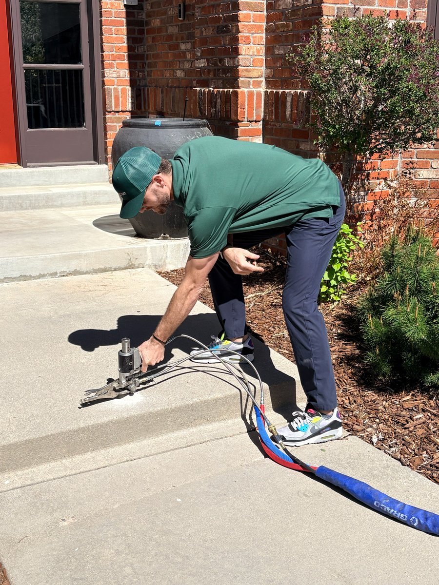 Tradesmen in green shirt and cap using a concrete cutting tool on a sidewalk near brick building steps.