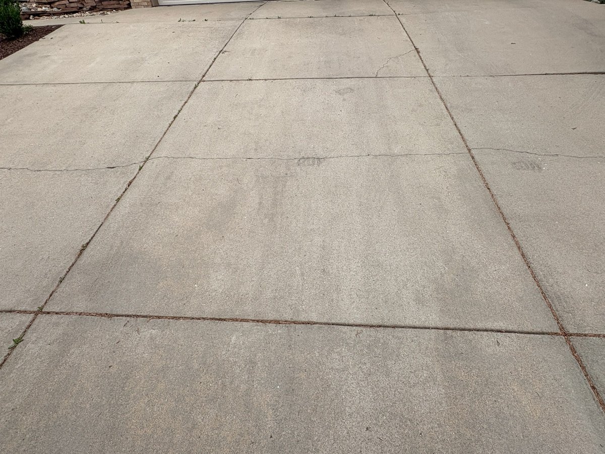 Concrete driveway with visible cracks and some dried pine needles in the seams.